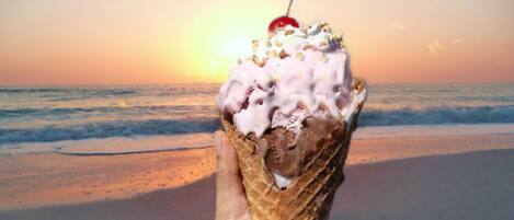 A hand holding an ice cream cone with a cherry on top, set against a beach backdrop with the ocean and a sunset.