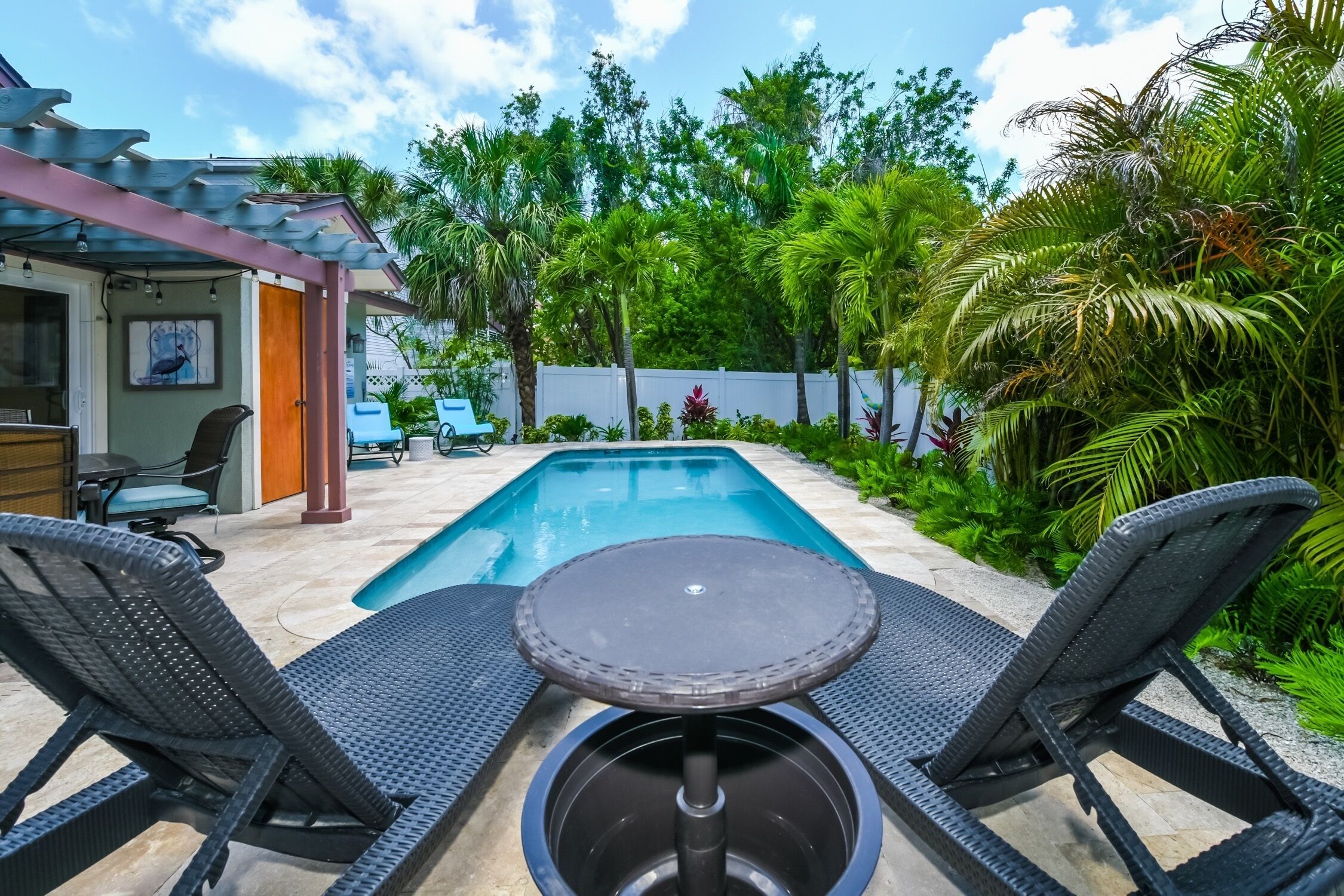 Un espace aménagé au bord de la piscine, avec deux chaises longues et une petite table, entouré d’une végétation luxuriante et d’une clôture blanche.