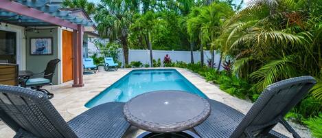 A poolside area with two lounge chairs and a small table, surrounded by lush greenery and a white fence.