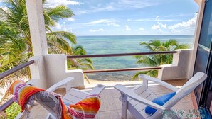 A balcony with white chairs and a colorful blanket, overlooking a beach with palm trees and the ocean. - Penthouse view of the Caribbean with AC, WiFi, easy beach access! (Akumal)