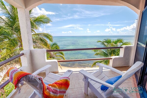 A balcony with white chairs and a colorful blanket, overlooking a beach with palm trees and the ocean.