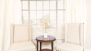Deux fauteuils placés face à face, avec une petite table ronde entre eux, un éventail décoratif posé sur la table, et une fenêtre avec des stores en arrière-plan.