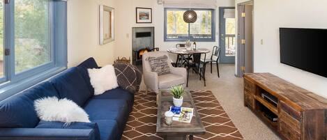 A living room with a blue sofa, a coffee table, and a TV. There is a fireplace, a dining area, and a large window.