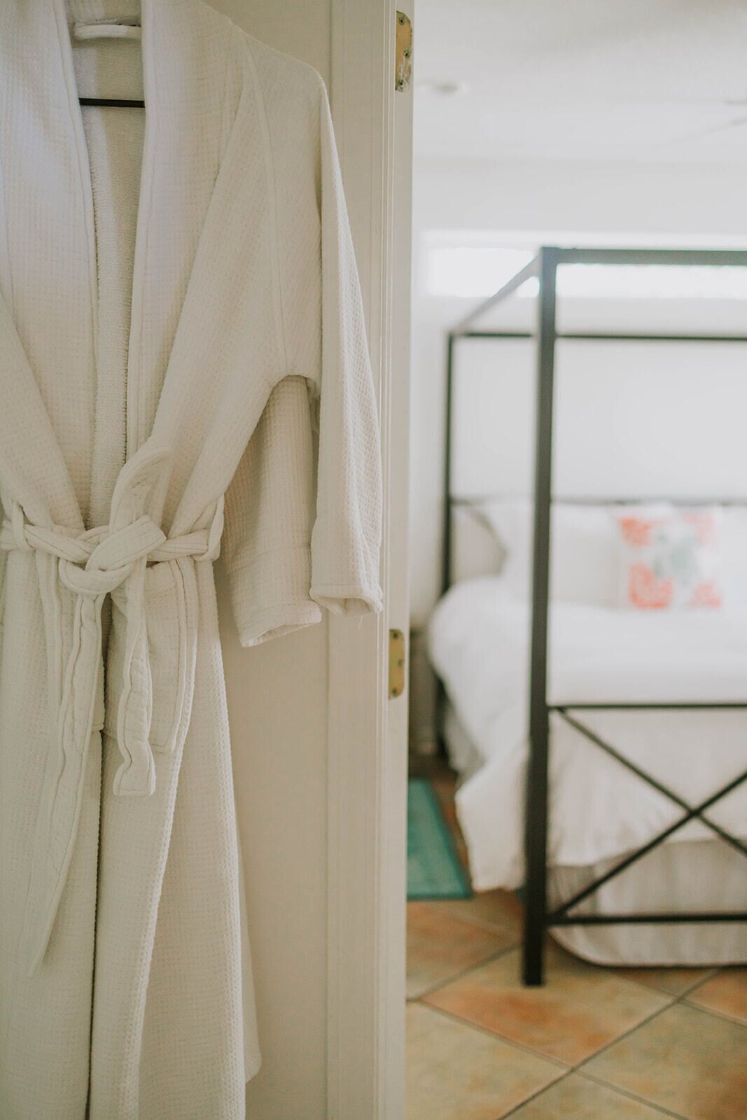 Un peignoir blanc est accroché à une porte, un lit superposé en métal est recouvert de draps blancs, et le sol est carrelé.