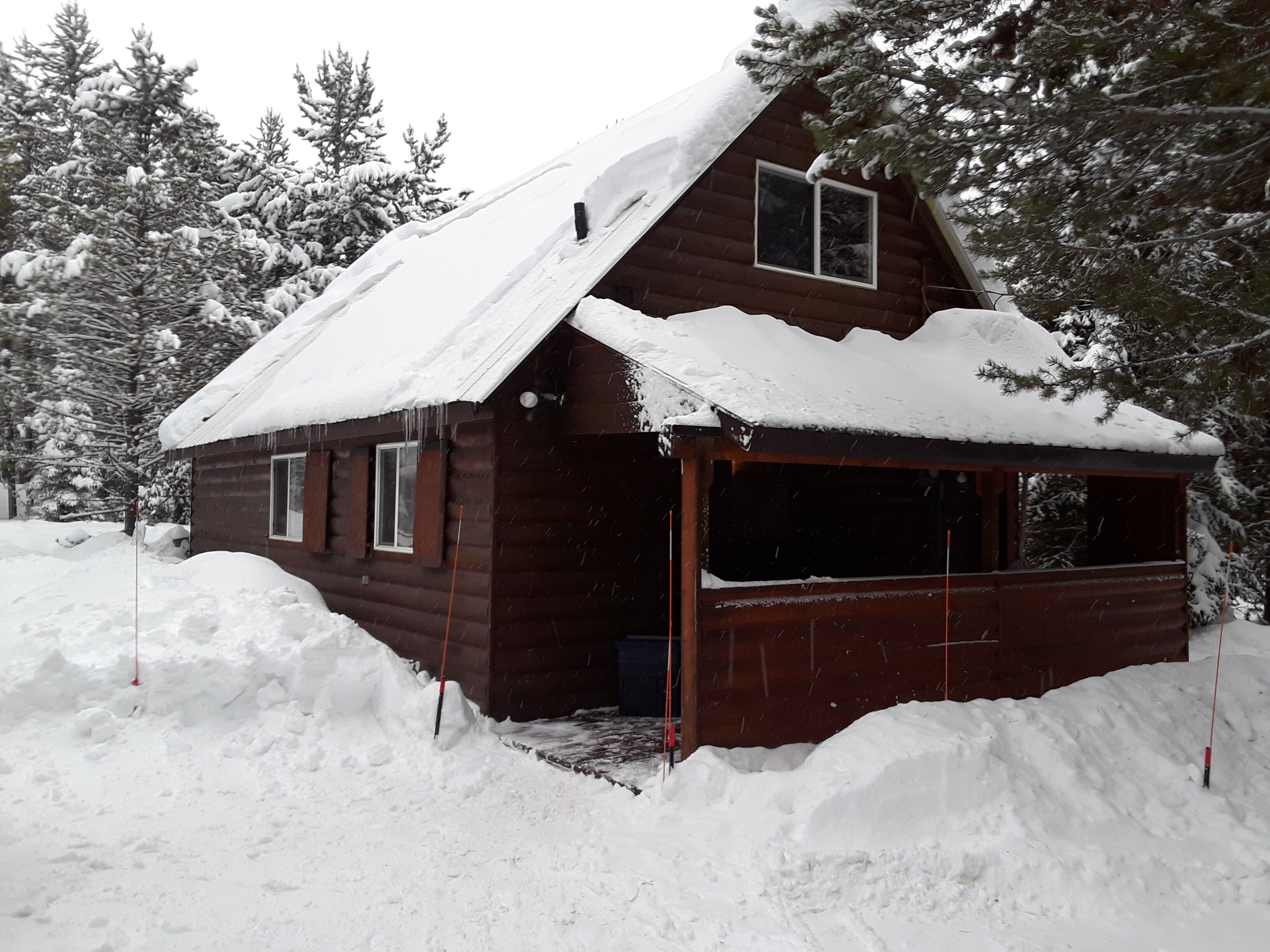 Una cabaña de madera con techo y ventanas cubiertas de nieve, rodeada de nieve y árboles.