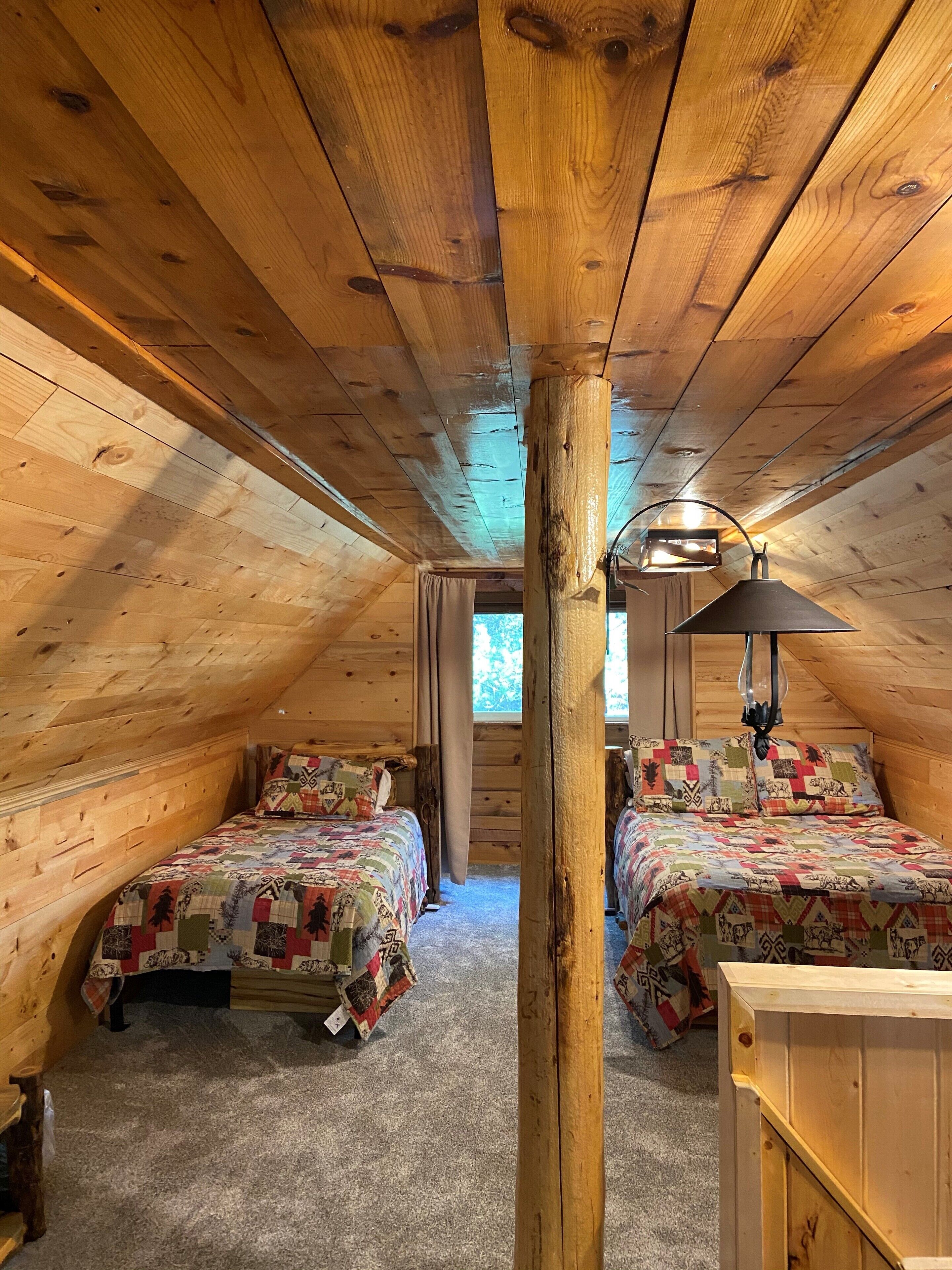 A cozy attic bedroom with two beds, wooden beams, a ceiling lamp, and a view of the outdoors.