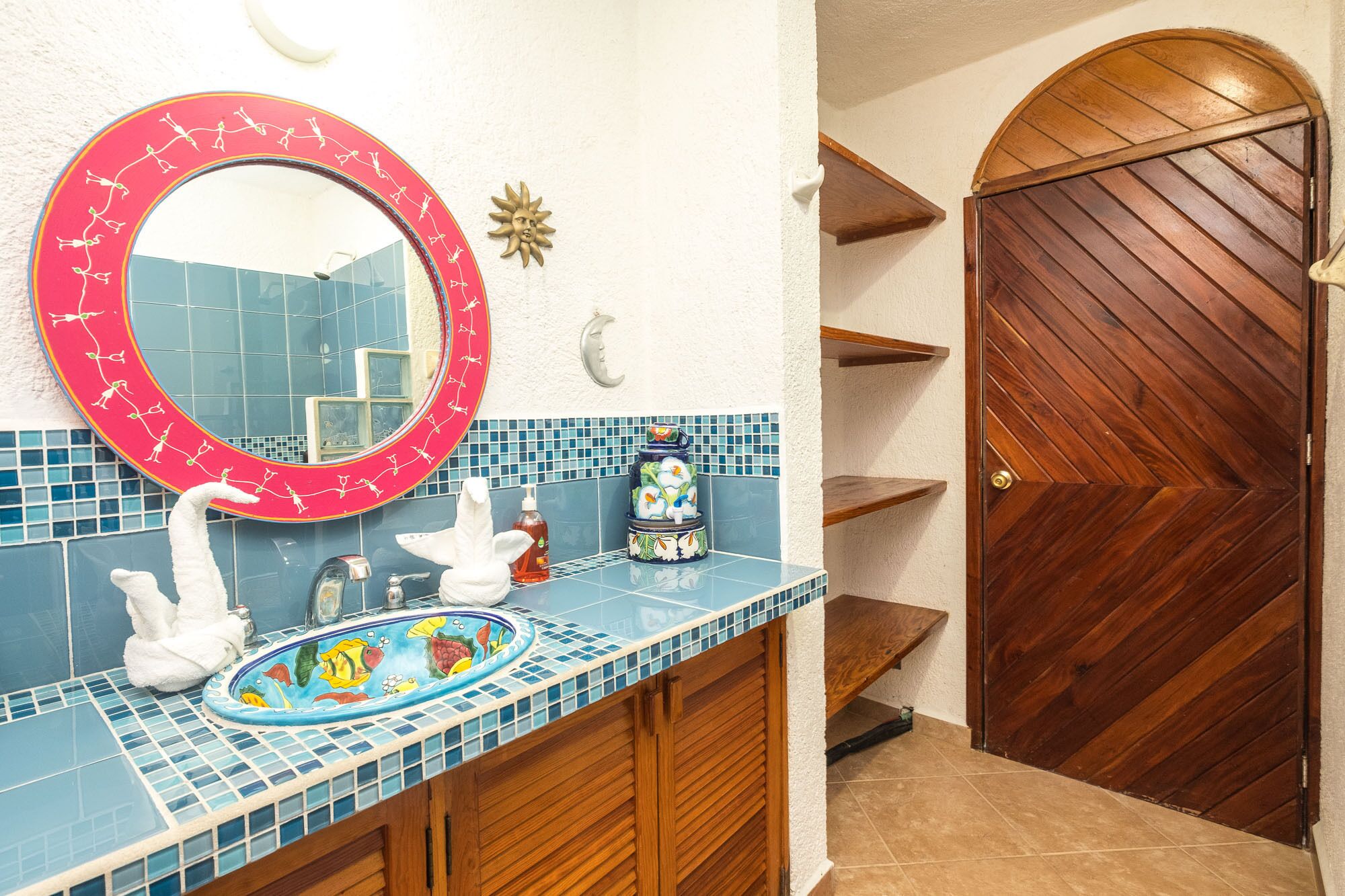 Une salle de bain avec un miroir rond, un lavabo dont le pare-éclaboussures est décoré de mosaïques et une porte en bois.