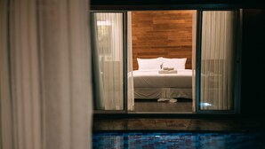 Une chambre avec une tête de lit en bois, un lit recouvert de draps blancs et une vue sur une piscine.