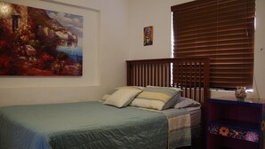 Une chambre à coucher avec un lit, un tableau, une tête de lit en bois, une fenêtre avec des stores et une étagère colorée.