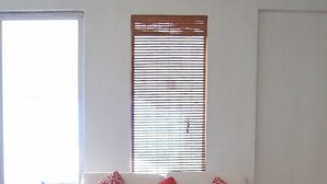 Un canapé blanc avec trois coussins rouges, un plancher en bois, un tapis blanc et une fenêtre avec des stores.
