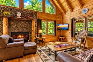 A cozy cabin interior with a stone fireplace, leather sofas, a television, and a large window offering a view of the outdoors.