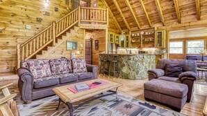A log cabin interior with a wooden staircase, a sofa, a coffee table, and a stone fireplace. - Glori's Retreat - $560 of FREE  tickets for each paid day (Pigeon Forge)