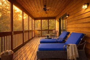 A covered porch with wicker lounge chairs, a ceiling fan, and a view of trees through large windows.