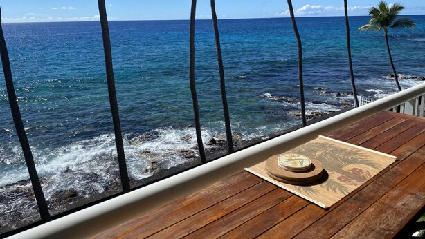 Une terrasse en bois avec une table en bois et un joli sous-verre décoratif, donnant sur une plage bordée de palmiers et sur l’océan.