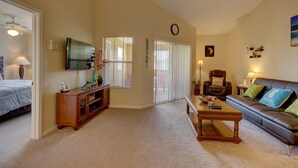 Ein Wohnzimmer mit Sofa, Couchtisch und Fernseher. Eine Tür führt in einen anderen Raum, und an der Wand hängt eine Uhr.