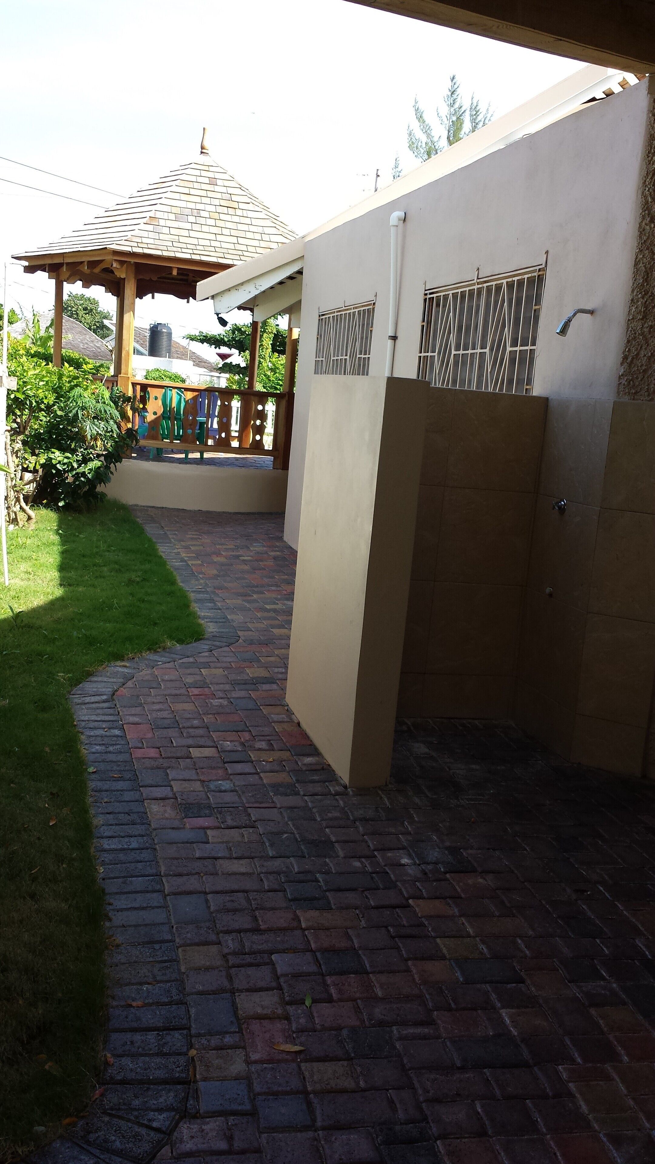 Un chemin en briques mène à un patio couvert agrémenté d’une pergola, d’une citerne et d’un mur avec des fenêtres grillagées.