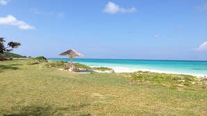 Ein Strand mit einem strohgedeckten Sonnenschirm und einem Tisch, umgeben von Grünflächen und freiem Blick auf den Ozean.