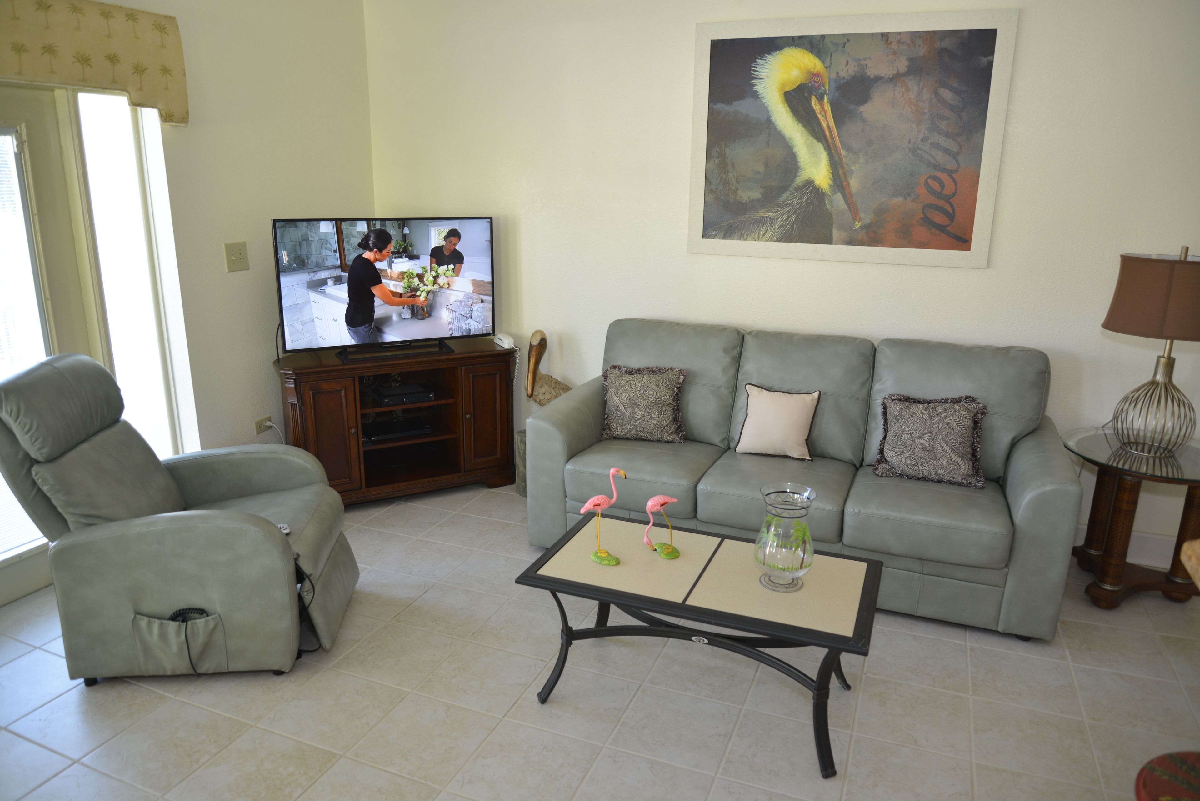 A living room with a grey sofa, a recliner, a coffee table with flamingo figurines, a television, and a painting of a bird.