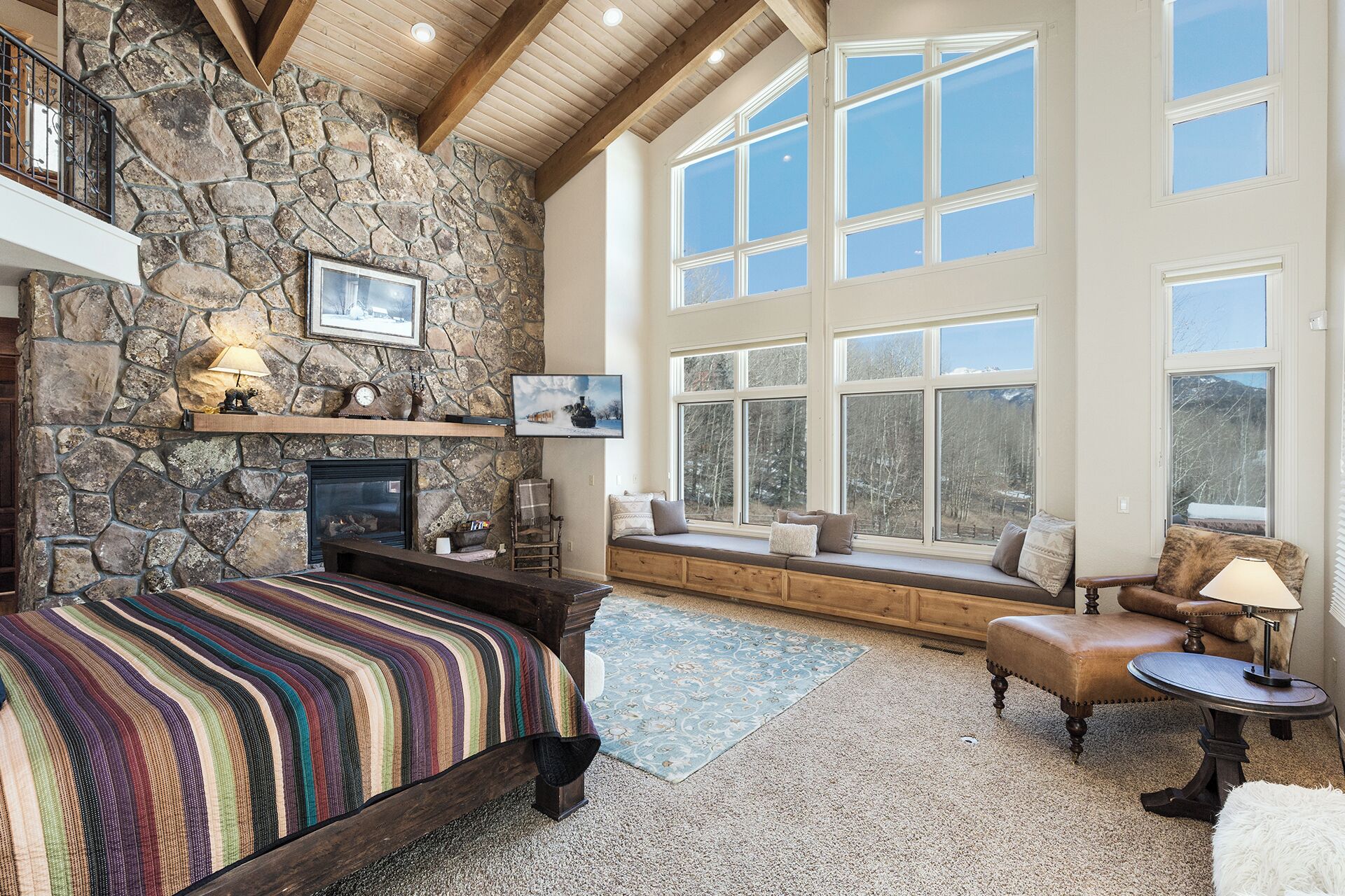 A cozy room with a stone fireplace, large windows, a comfortable seating area, and a bed with a colorful striped blanket.