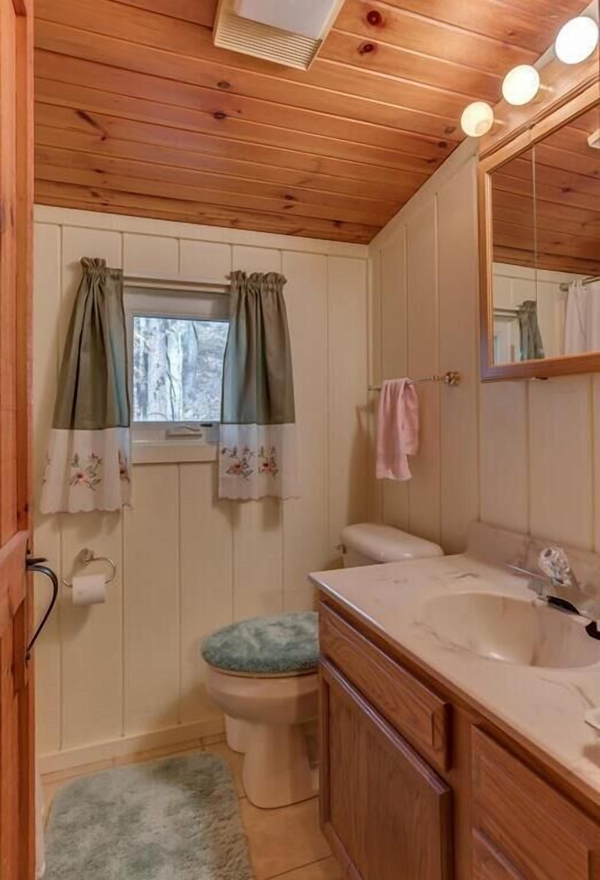 Un baño pequeño y funcional con paredes de paneles de madera, un inodoro con tapa azul, un lavamanos con espejo y una ventana con cortinas.