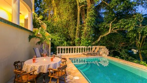 Uma área à beira da piscina com uma mesa redonda e cadeiras, rodeada por vegetação exuberante e um portão decorativo.