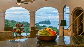 Uma área de refeições com vista para o mar, um ventilador de teto e uma tigela de frutas na mesa.
