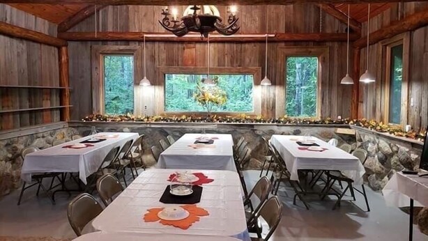 A rustic dining hall with wooden walls, multiple tables set for dining, and large windows with a forest view. - Full Log Lodge 3 night minimum, 2 night  minimum  October- April, Pet Friendly  (Lakewood)