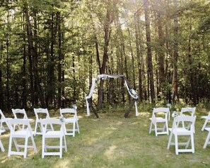 Ceremonia de boda al aire libre con sillas blancas dispuestas en filas, un arco decorado y un fondo de bosque.