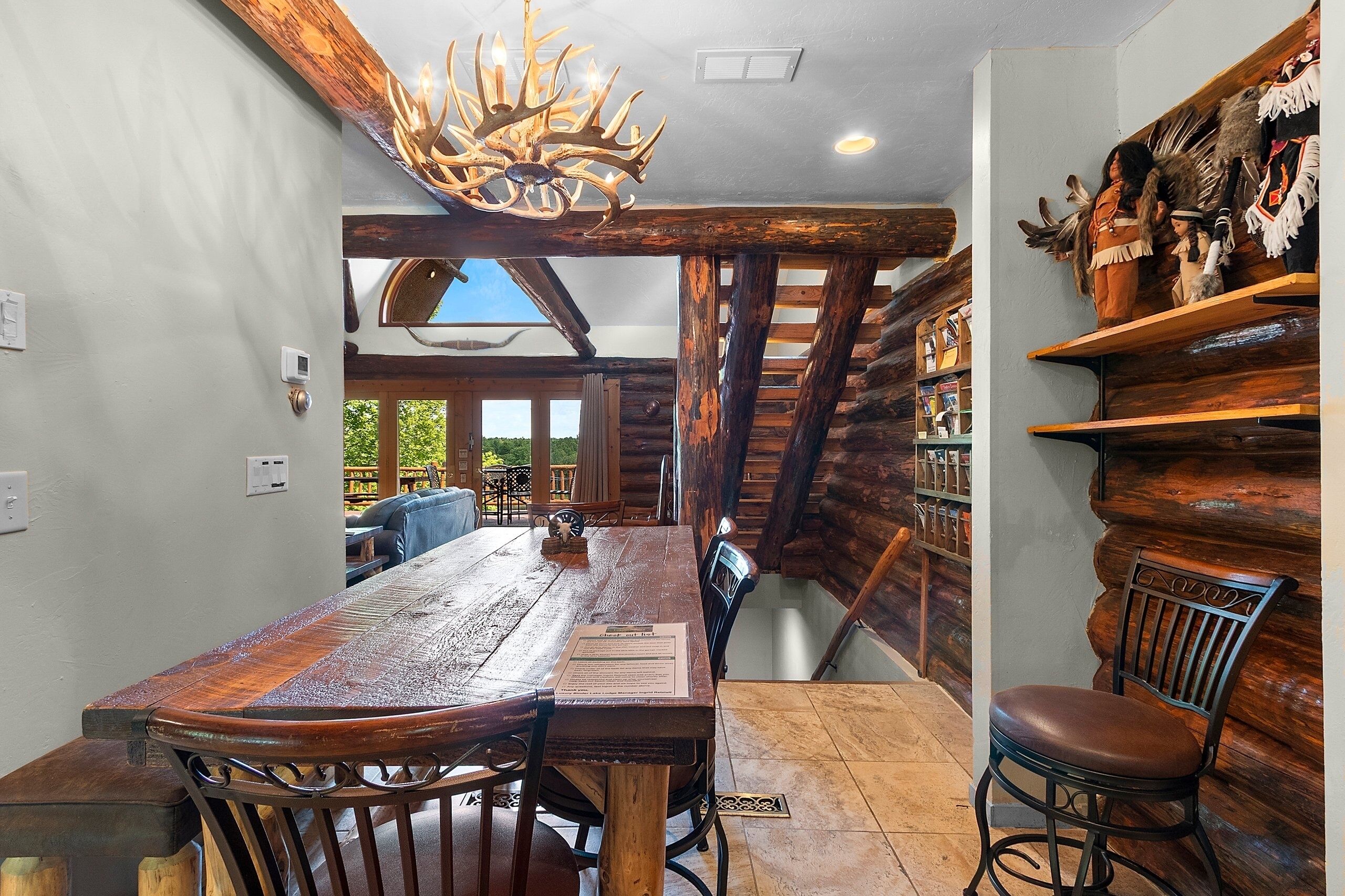 A rustic interior with a wooden dining table, chairs, a chandelier, and a bookshelf with decorative items.