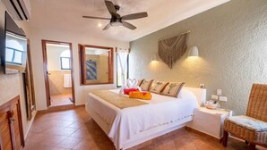 Une chambre d’hôtel avec un grand lit, un ventilateur de plafond, une petite table avec une chaise, et une vue sur la salle de bain.