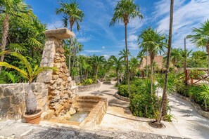 Un complex turístic tropical amb una font de pedra, palmeres i un edifici amb teulada de palla.