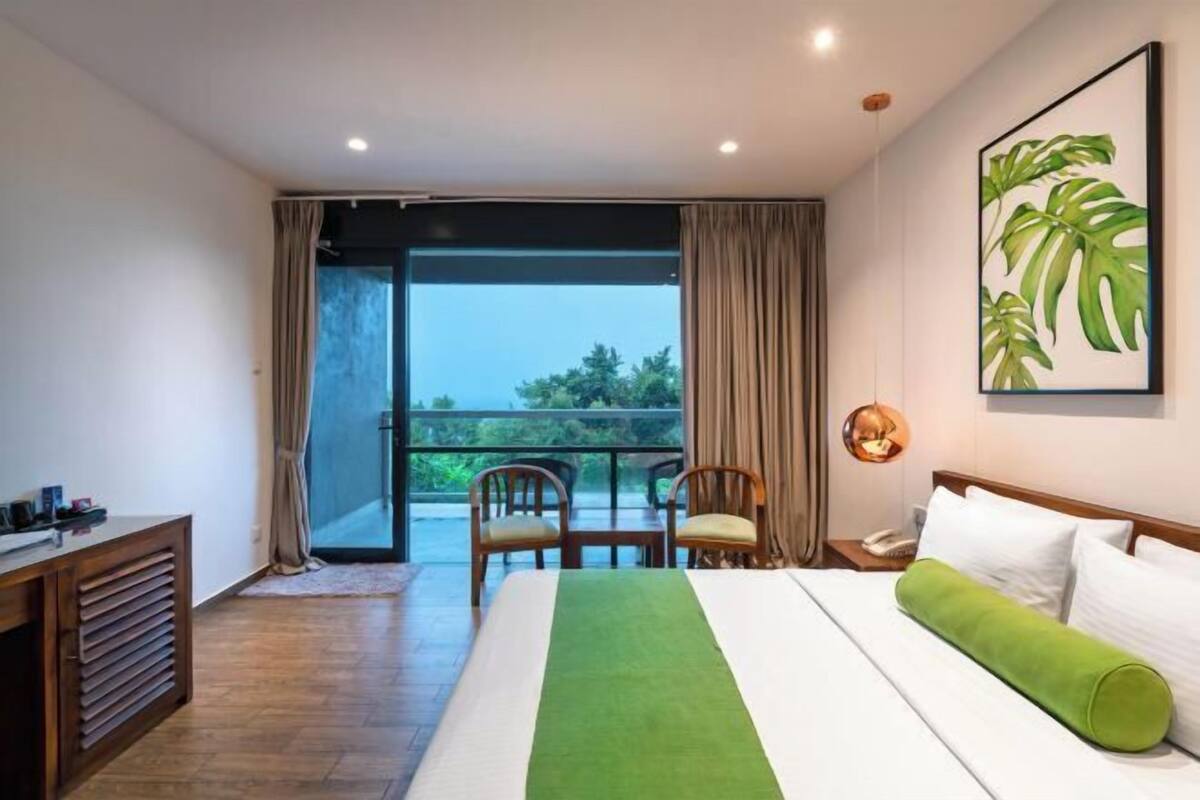 A hotel room with a bed, a desk, chairs, a balcony with curtains, and a large window.