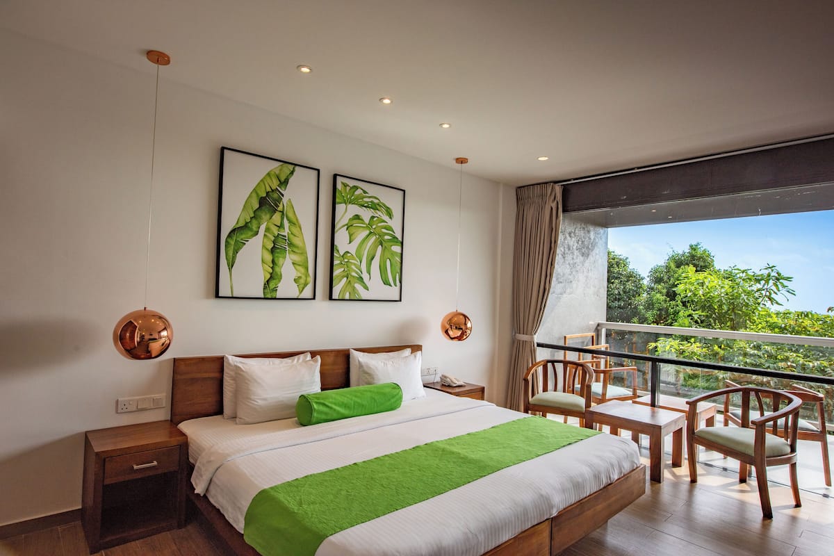 A modern hotel room with a large bed, a balcony with wooden chairs, and two framed plant artworks on the wall.