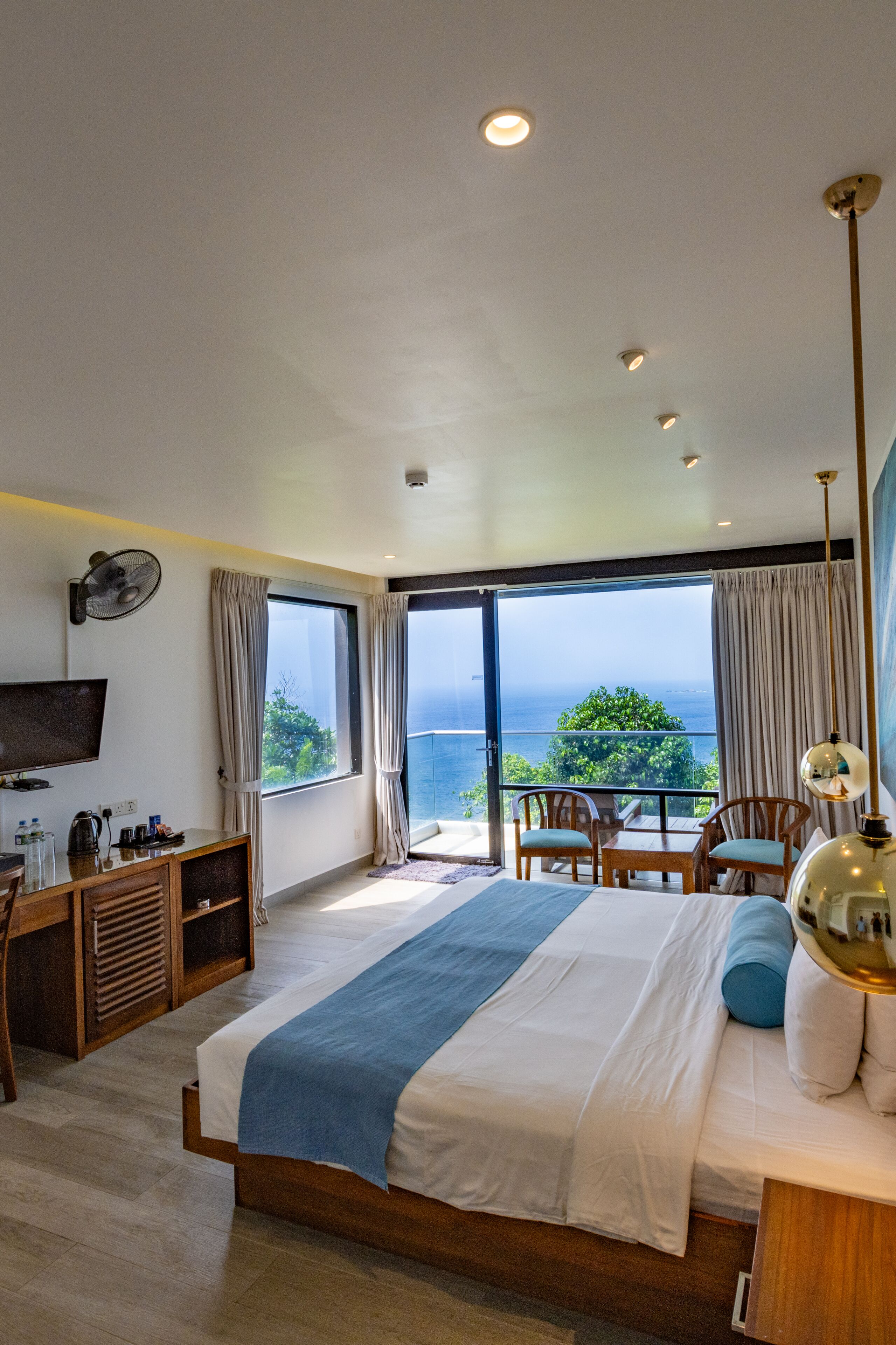 A modern hotel room with a large bed, a desk with a flat-screen TV, a seating area with a table and chairs, and a balcony with a view of the ocean.