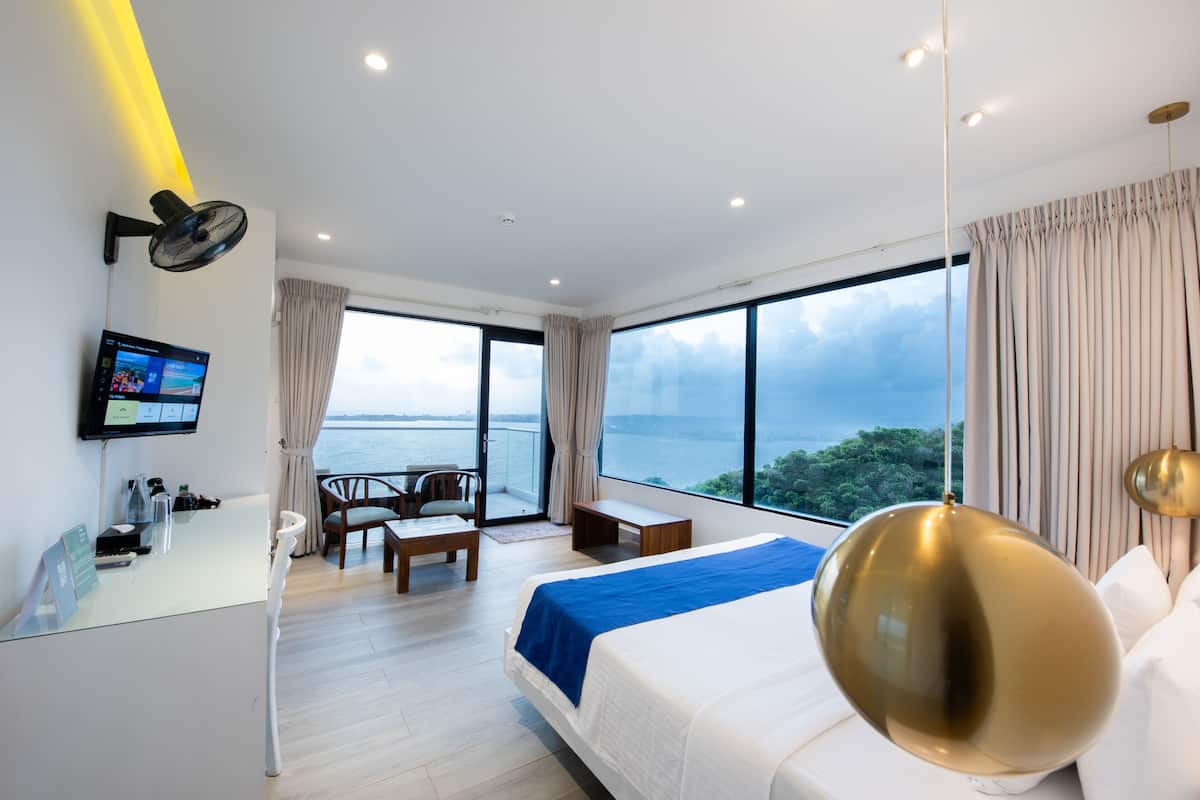 A modern hotel room with a large bed, a desk with a computer, a TV, and a balcony with a view of the sea.