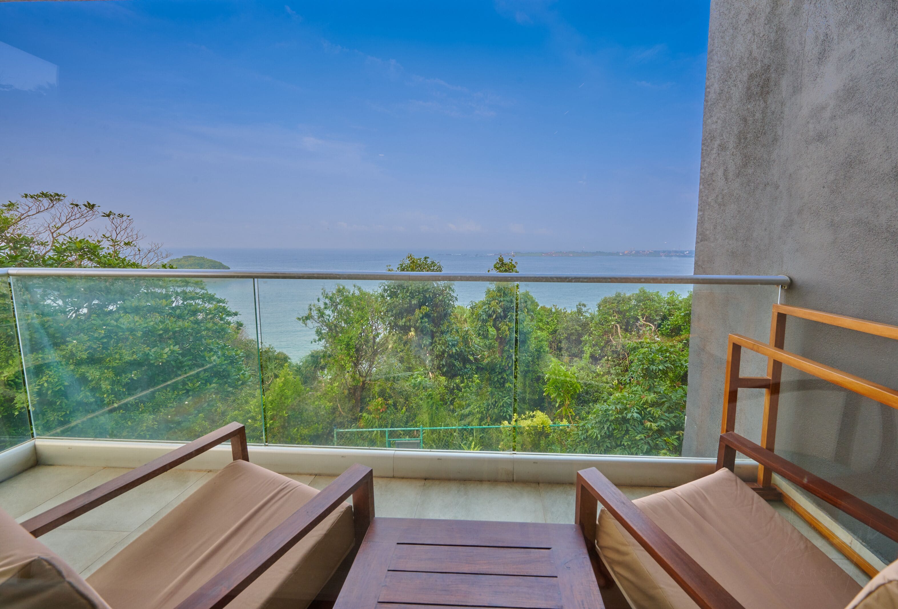 A balcony with two wooden chairs and a small table, overlooking a lush green landscape and the sea.
