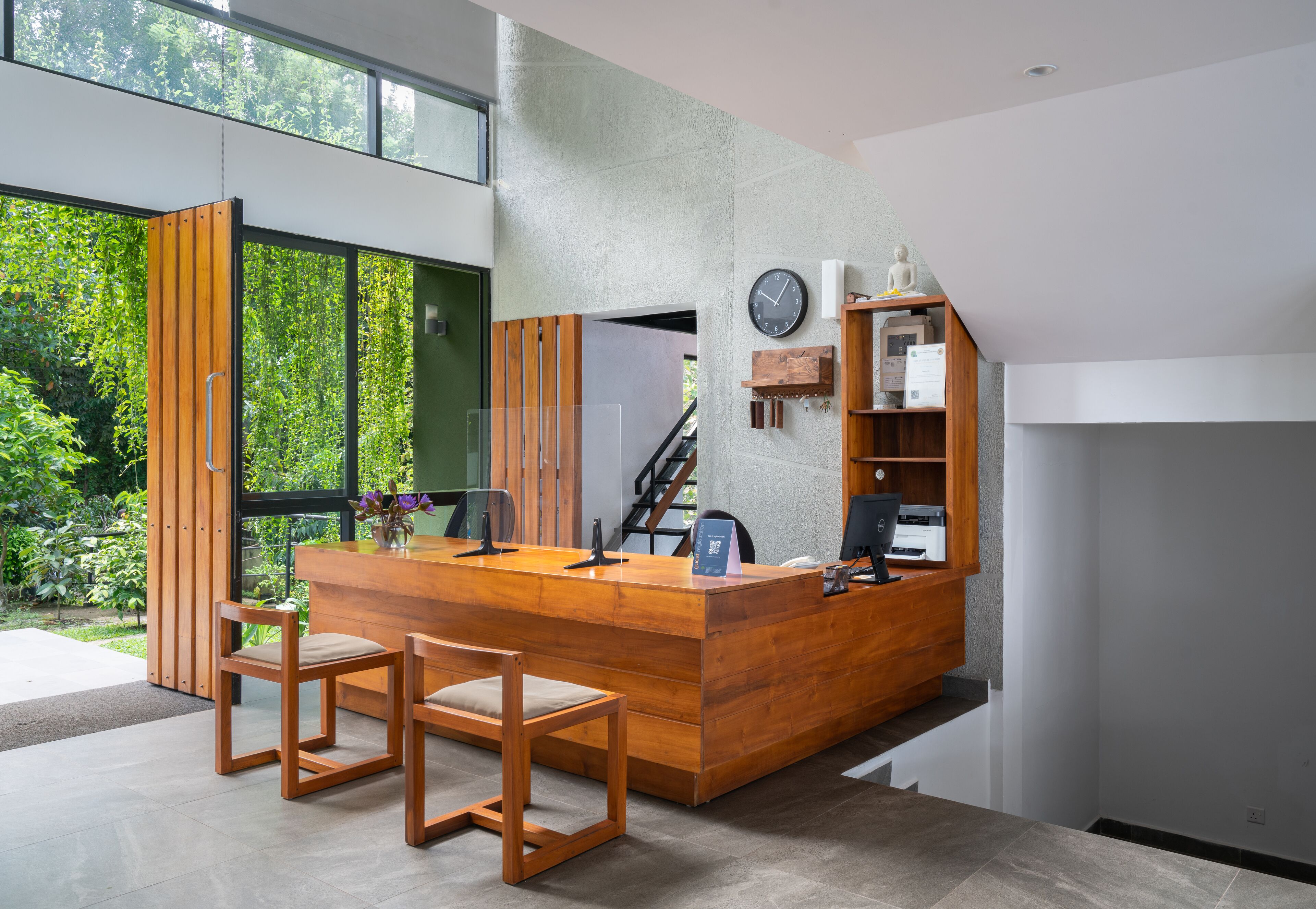 A modern reception desk with wooden chairs, a clock, and a view of greenery outside.