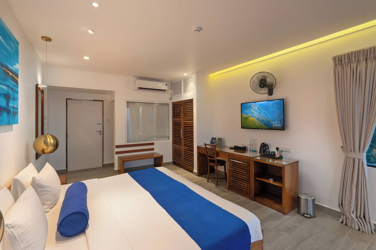 A modern hotel room with a large bed, a desk, a chair, a TV, and a small trash bin.