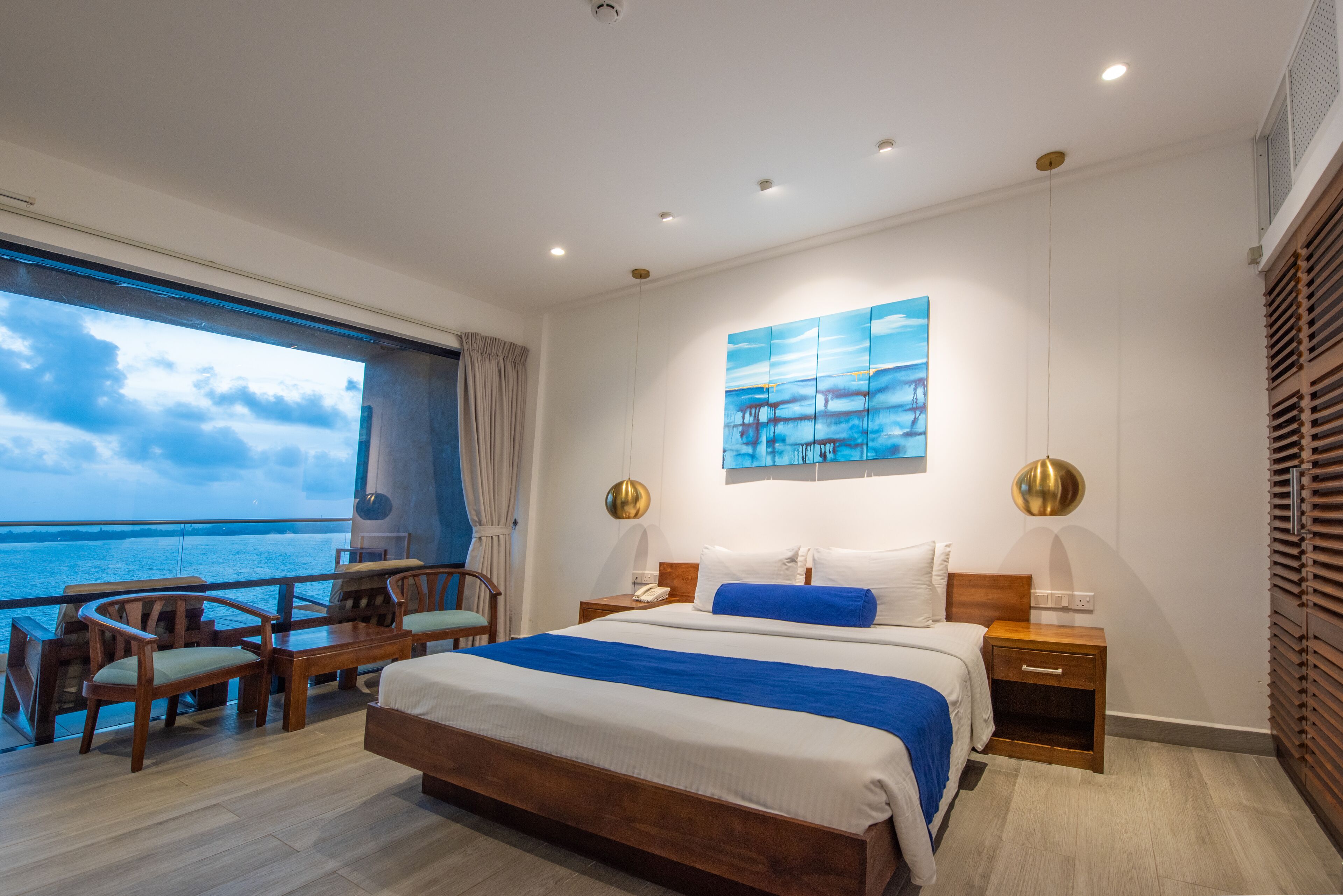 A modern hotel room with a large bed, wooden furniture, a view of the sea, and a painting on the wall.