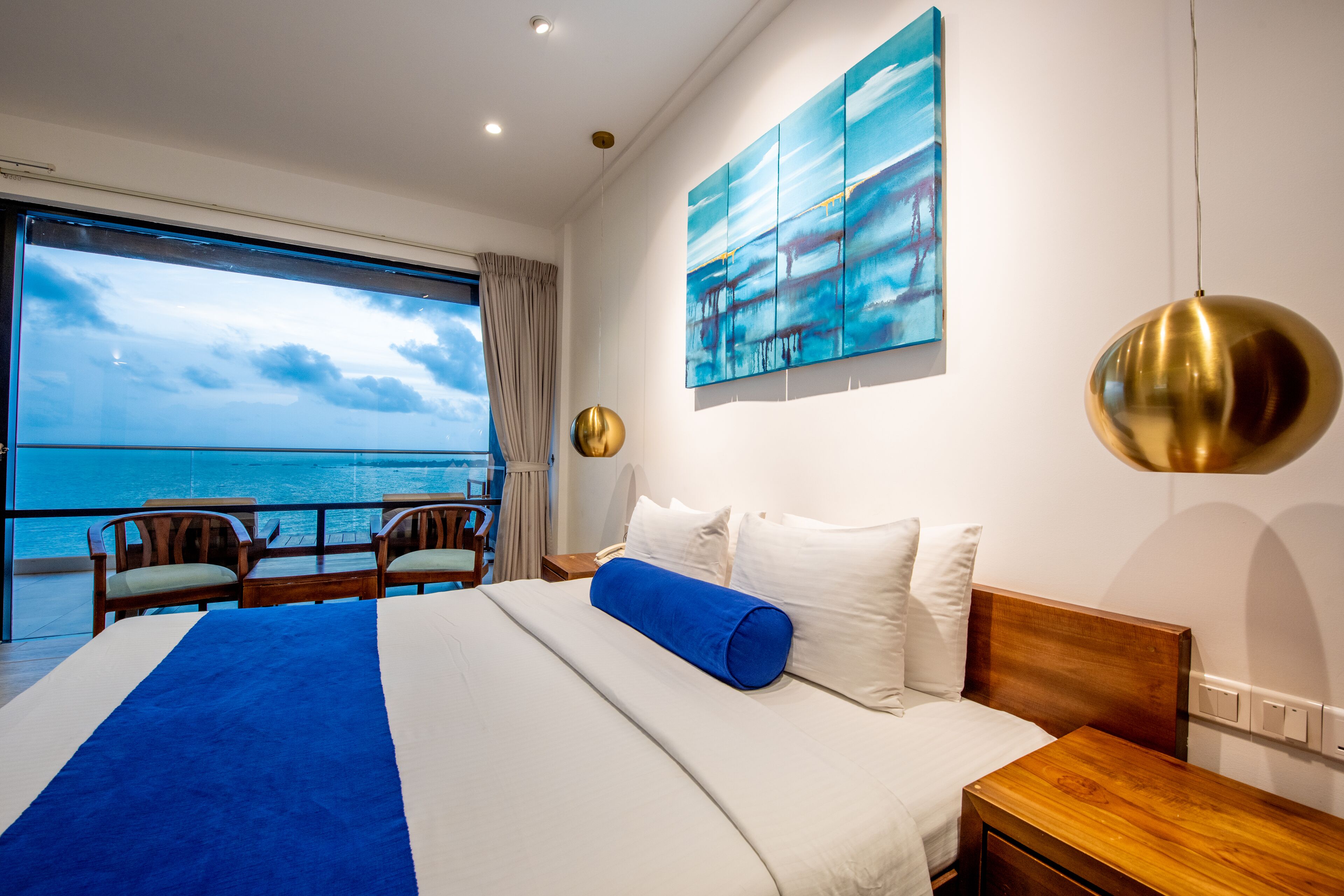 A hotel room with a large bed, a view of the sea, a painting on the wall, and a pendant light.