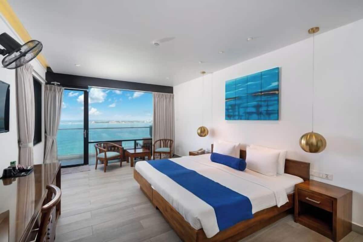 A hotel room with a bed, a desk, a chair, a TV, and a view of the sea.