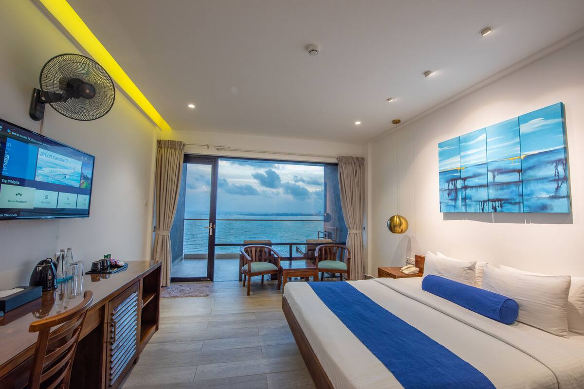 A hotel room with a bed, a desk with a computer, a fan, a TV, a balcony with a view of the sea, and a painting on the wall.
