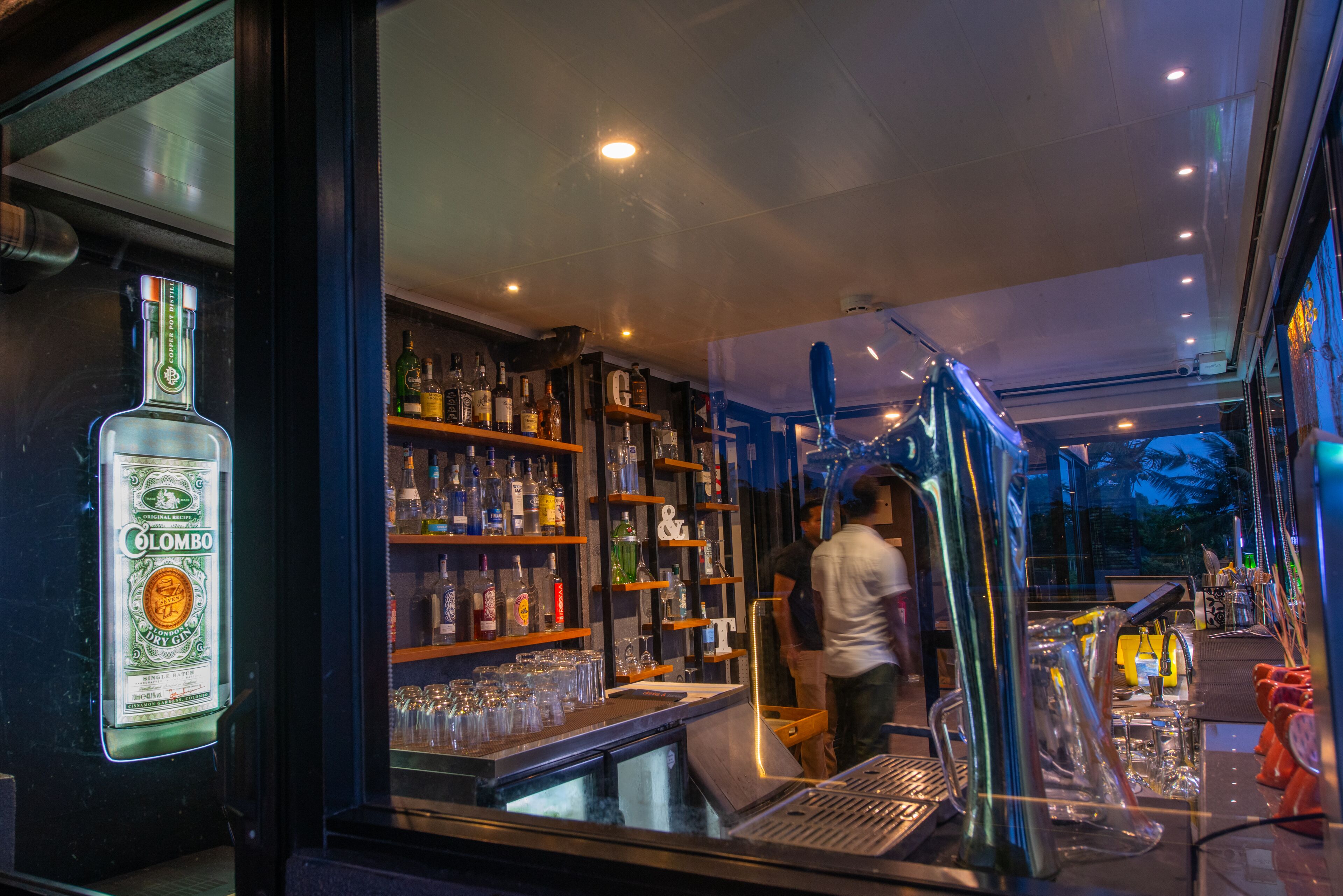 A bar area with shelves of bottles and glasses, a bar counter, and a person standing near the bar.