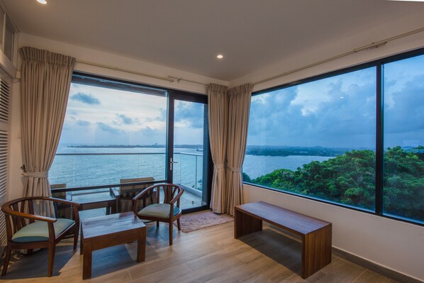 A room with a large window overlooking a body of water and greenery. There is a wooden bench, two chairs, and a small table. - Agnus Unawatuna (Unawatuna)