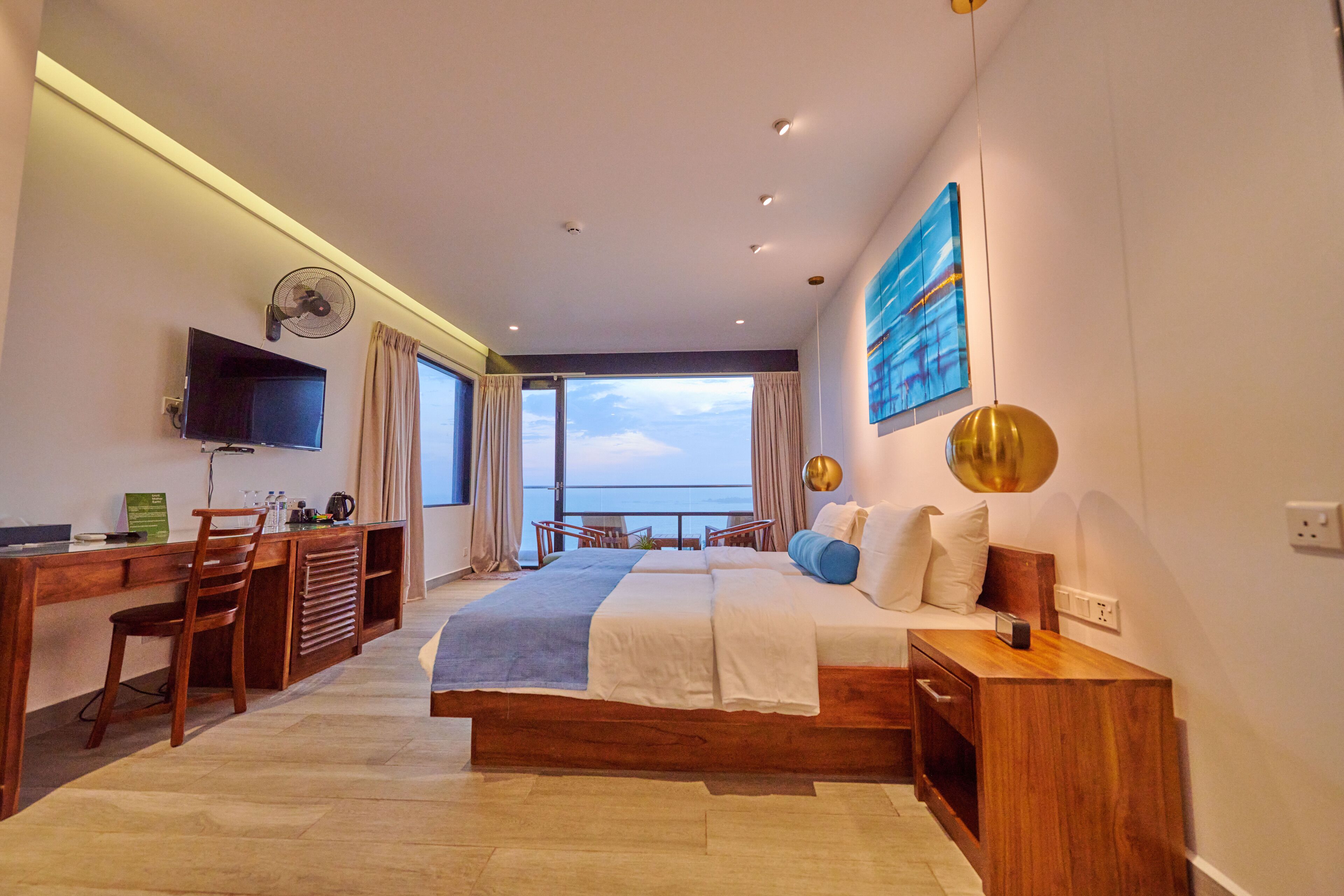 A modern hotel room with a large bed, a desk with a chair, a television, and a view of the sea.