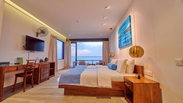 A modern hotel room with a large bed, a desk with a chair, a television, and a view of the sea.