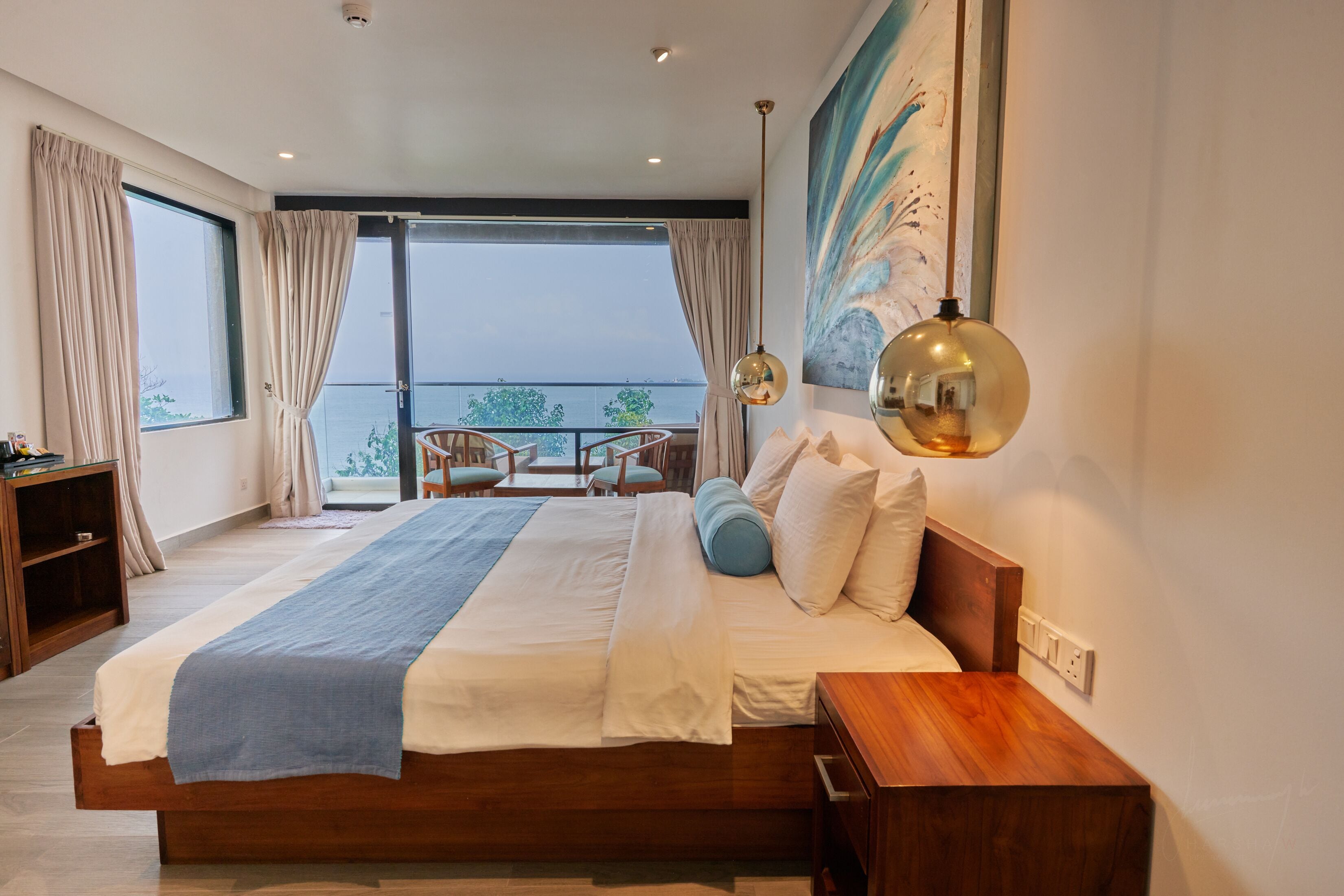 A modern hotel room with a large bed, a balcony with a view, a dining area, and a painting on the wall.