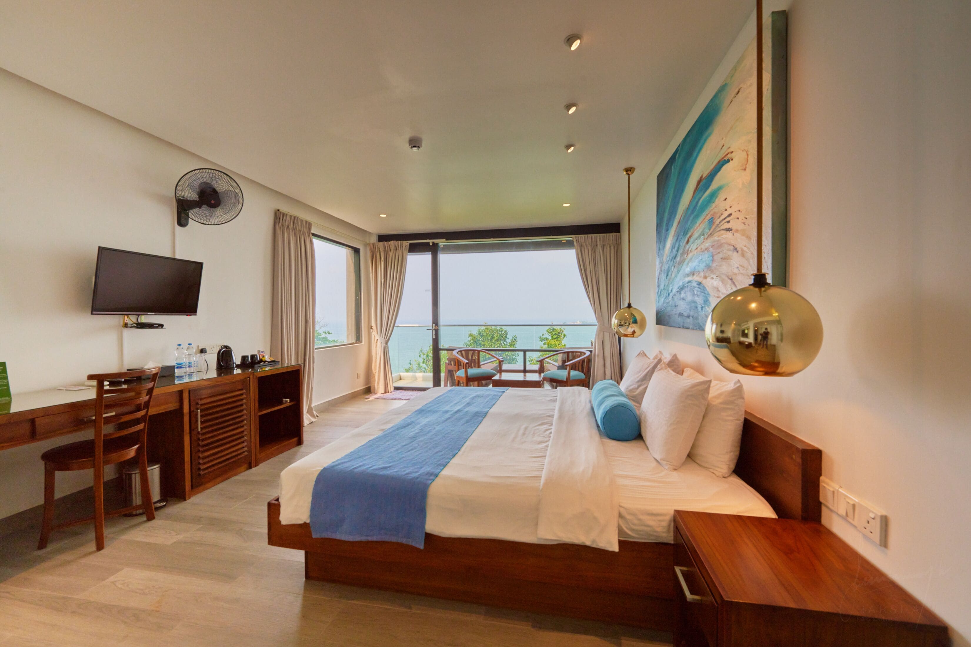 A modern hotel room with a large bed, a desk with a flat-screen TV, a balcony with a view, and a large painting on the wall.