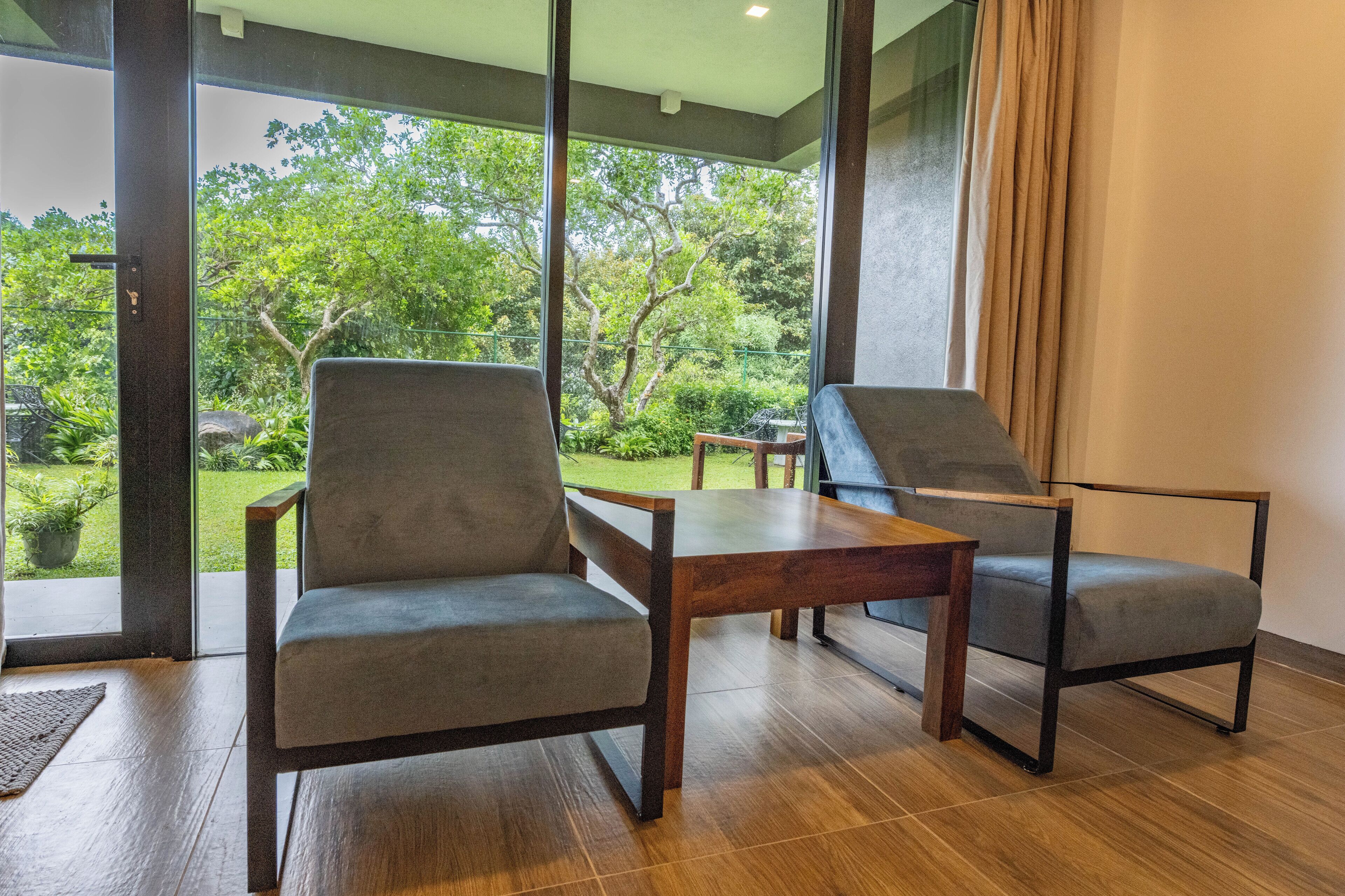 Una habitación moderna con dos sillones grises y una mesa de madera, ventanas amplias con vistas al exterior, lleno de vegetación.