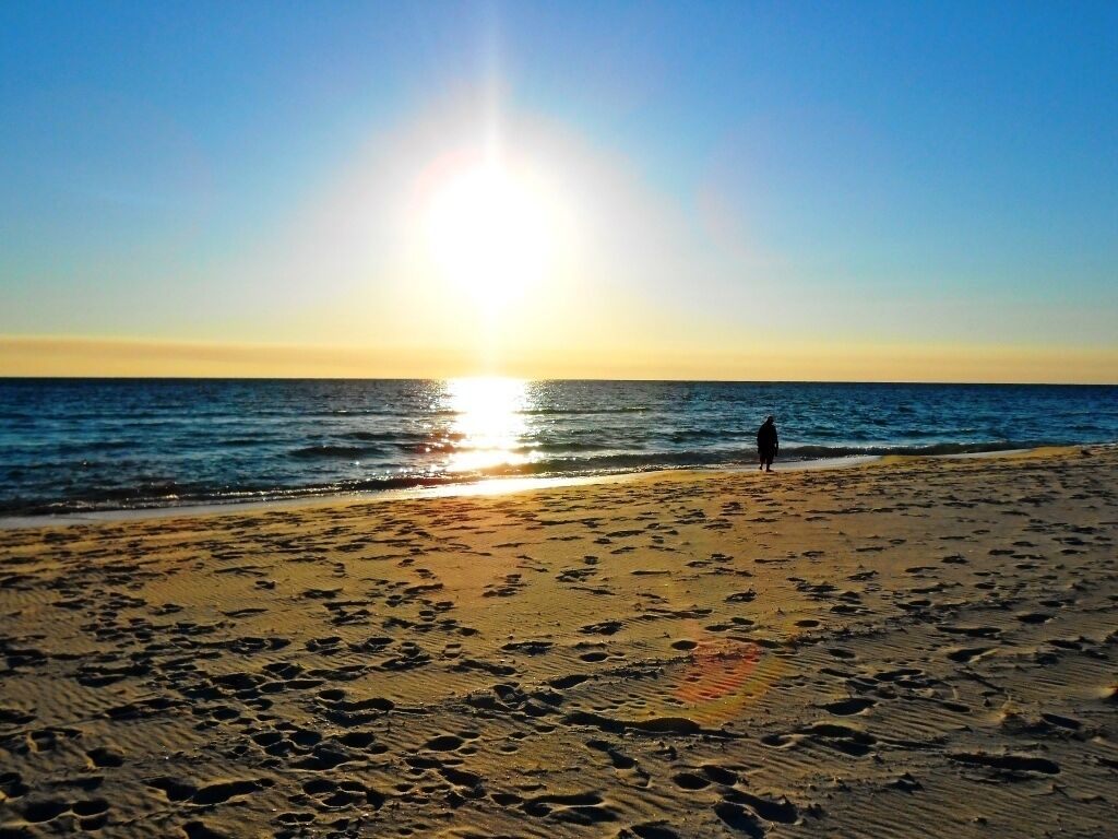 Una playa con huellas que conducen al agua, una persona de pie cerca de la orilla y el sol poniéndose en el horizonte.